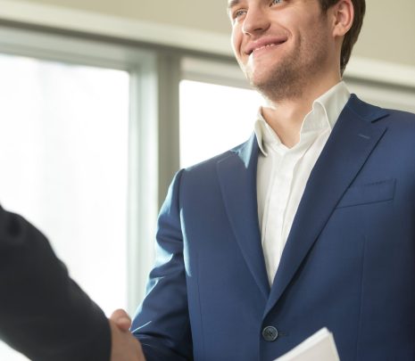 Confident smiling businessman with documents in hand handshaking with partner at business meeting in office. Presentable financial consultant welcoming client, congratulating investor with good deal